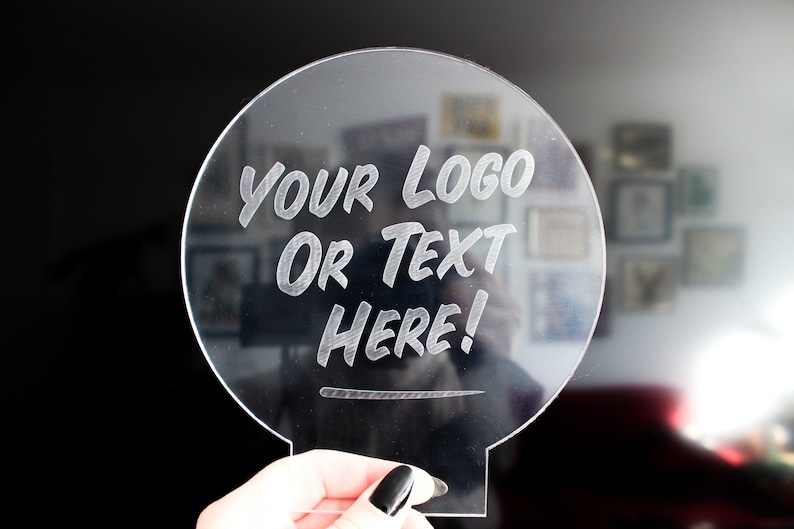 May include: Clear acrylic sign with a circular top and a rectangular base. The sign features the engraved text "YOUR LOGO OR TEXT HERE!". The background is blurred, with a dark area on the left and a wall with framed pictures on the right.
