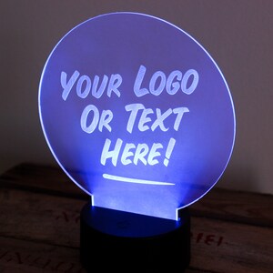 May include: A glowing blue LED sign with the text "YOUR LOGO OR TEXT HERE!" in white. The sign is circular and mounted on a black base. The background is a dark wooden surface.