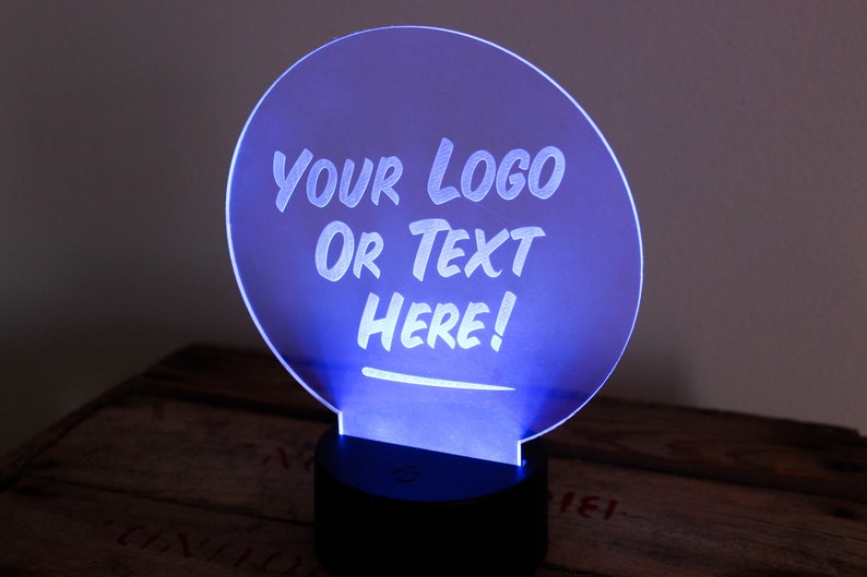 May include: A glowing blue LED sign with the text "YOUR LOGO OR TEXT HERE!" in white. The sign is circular and mounted on a black base. The background is a dark wooden surface.
