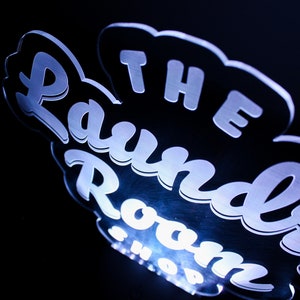 May include: A glowing sign with the words "The Laundry Room Shop" in a stylized font. The sign is white with a blue glow against a black background. The design is in a cloud-like shape.