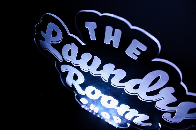 May include: A glowing sign with the words "The Laundry Room Shop" in a stylized font. The sign is white with a blue glow against a black background. The design is in a cloud-like shape.