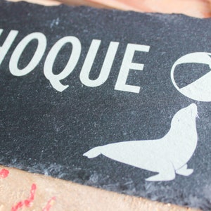 Puede incluir: Un letrero de pizarra negra con texto blanco que dice "PHOQUE" y una silueta blanca de una foca equilibrando una pelota de playa en su nariz.