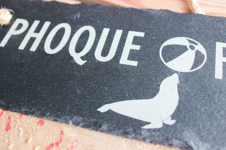Puede incluir: Un letrero de pizarra negra con texto blanco que dice "PHOQUE" y una silueta blanca de una foca equilibrando una pelota de playa en su nariz.