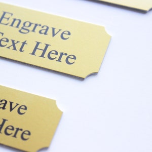 K&ouml;nnte beinhalten: Zwei goldfarbene Metallplaketten mit abgerundeten Ecken. Die obere Plakette tr&auml;gt den Text "We Engrave Your Text Here". Die untere Plakette tr&auml;gt den Text "Engrave Text Here".