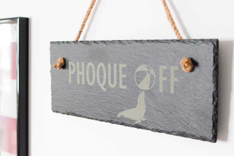 Puede incluir: Un letrero de pizarra negra con el texto "PHOOQUE OFF" en letras blancas. Una silueta blanca de una foca equilibrando una pelota de playa est&aacute; debajo del texto. El letrero est&aacute; colgado de una cuerda con nudos en cada extremo.