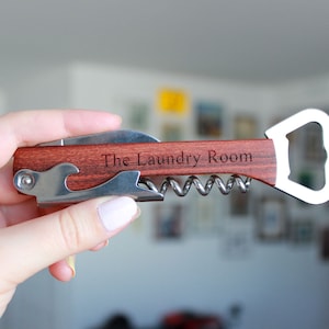 Könnte beinhalten: Ein Multi-Tool mit Holzgriff und Edelstahlkomponenten. Verfügt über einen Flaschenöffner, einen Korkenzieher und ein Messer. Der Text "The Laundry Room" ist in das Holz graviert. Ein praktisches und stilvolles Bar-Accessoire.