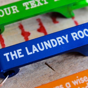 Op de afbeelding: Drie flessenopeners van verschillende gekleurde metalen met tekst. De bovenste opener is groen met de tekst "YOUR TEXT". De middelste opener is blauw met de tekst "THE LAUNDRY ROOM". De onderste opener is oranje met de tekst "as a wise man said beer".