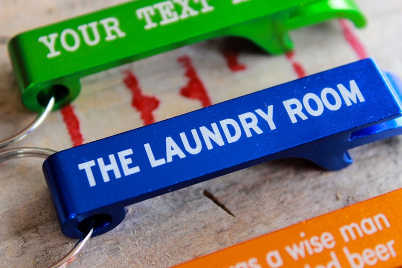 Op de afbeelding: Drie flessenopeners van verschillende gekleurde metalen met tekst. De bovenste opener is groen met de tekst "YOUR TEXT". De middelste opener is blauw met de tekst "THE LAUNDRY ROOM". De onderste opener is oranje met de tekst "as a wise man said beer".