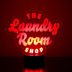 May include: A glowing red and yellow LED sign that reads "The Laundry Room Shop." The sign is in a cursive font and sits on a black base, with a red background.