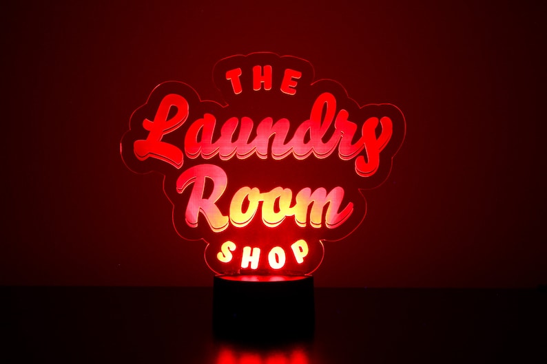 May include: A glowing red and yellow LED sign that reads "The Laundry Room Shop." The sign is in a cursive font and sits on a black base, with a red background.