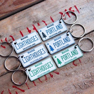 May include: Keychains with license plate designs. The keychains feature the words "MAINE LIGHTHOUSES VACATIONLAND" and "MAINE PORTLAND VACATIONLAND" in white text on a blue or green background. Each keychain has a silver key ring.