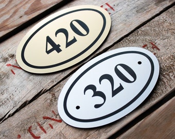 Könnte beinhalten: Zwei ovale Hausnummernschilder. Eines ist golden mit schwarzen Zahlen "420" und das andere ist silberfarben mit schwarzen Zahlen ".320".