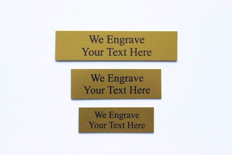 K&ouml;nnte beinhalten: Drei goldfarbene Metallplaketten mit dem Text "We Engrave Your Text Here" auf jeder.