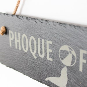 Puede incluir: Un letrero de pizarra negra con el texto "PHOOQUE OFF" en letras blancas. Una silueta blanca de una foca sosteniendo una pelota de playa est&aacute; al lado del texto. El letrero est&aacute; colgado de una cuerda.