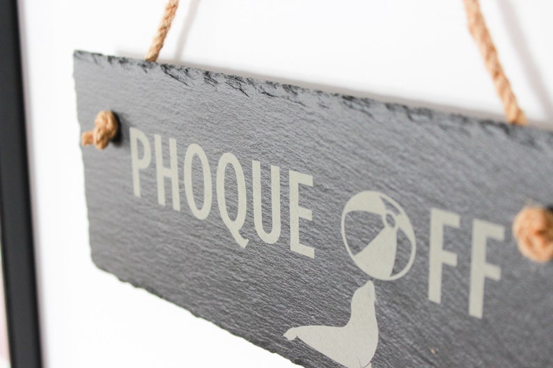 Puede incluir: Un letrero de pizarra negra con el texto "PHOOQUE OFF" en letras blancas. Una silueta blanca de una foca sosteniendo una pelota de playa est&aacute; al lado del texto. El letrero est&aacute; colgado de una cuerda.