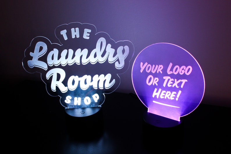 May include: Two illuminated LED signs. One sign is shaped like a cloud with the words "THE Laundry Room SHOP" in white. The other is a purple circle with the text "YOUR LOGO OR TEXT HERE!" in white.