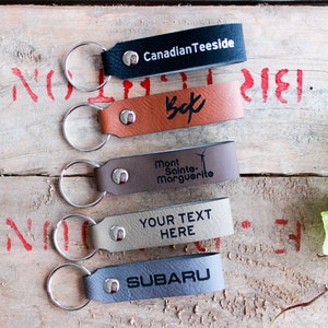 May include: Five leather keychains with metal rings. The keychains are brown, grey, and black. The keychains are engraved with the text "Canadian Teeside", "Bek", "Mont Sainte-Marguerite", "YOUR TEXT HERE", and "SUBARU".