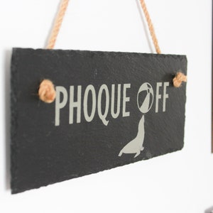 Puede incluir: Un letrero de pizarra negra con el texto "PHOCQUE OFF" en letras blancas. Debajo del texto hay una silueta blanca de una foca con una pelota de playa. El letrero est&aacute; colgado de un trozo de cuerda.
