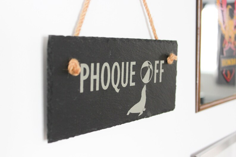 Puede incluir: Un letrero de pizarra negra con el texto "PHOCQUE OFF" en letras blancas. Debajo del texto hay una silueta blanca de una foca con una pelota de playa. El letrero est&aacute; colgado de un trozo de cuerda.