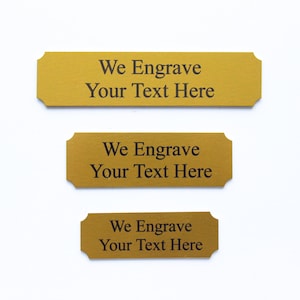 K&ouml;nnte beinhalten: Drei goldfarbene Metallplaketten mit abgerundeten Ecken und einer abgeschr&auml;gten Kante. Jede Plakette tr&auml;gt den Text "We Engrave Your Text Here", der in Schwarz graviert ist.