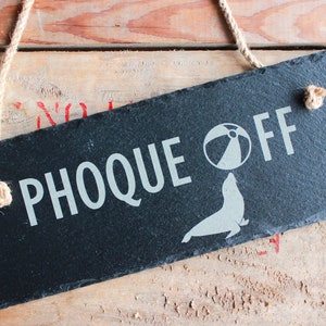 Puede incluir: Un letrero de pizarra negra con texto blanco que dice "Phoque Off" con un gr&aacute;fico de una foca equilibrando una pelota de playa en su nariz.