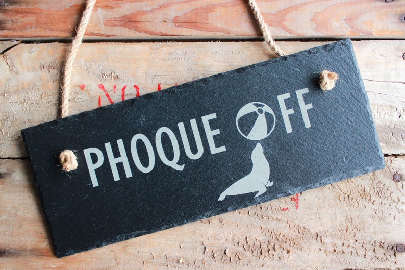 Puede incluir: Un letrero de pizarra negra con texto blanco que dice "Phoque Off" con un gr&aacute;fico de una foca equilibrando una pelota de playa en su nariz.