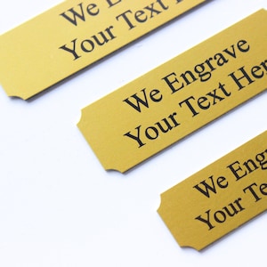 K&ouml;nnte beinhalten: Drei goldfarbene Metall-Namensschilder mit abgerundeten Ecken und gewellten Kanten. Jede Platte hat schwarzen Text mit der Aufschrift "We Engrave Your Text Here".
