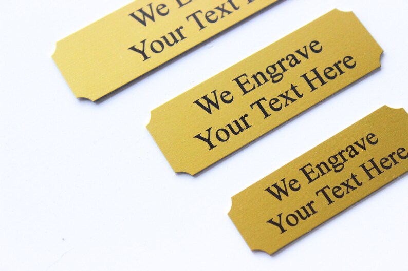 K&ouml;nnte beinhalten: Drei goldfarbene Metall-Namensschilder mit abgerundeten Ecken und gewellten Kanten. Jede Platte hat schwarzen Text mit der Aufschrift "We Engrave Your Text Here".