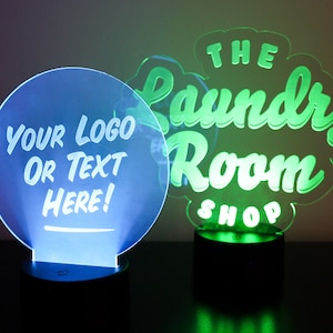 May include: Two illuminated LED signs. One is a blue circle with the text "YOUR LOGO OR TEXT HERE!". The other is a green sign that says "THE Laundry Room SHOP". Both signs are on black bases.