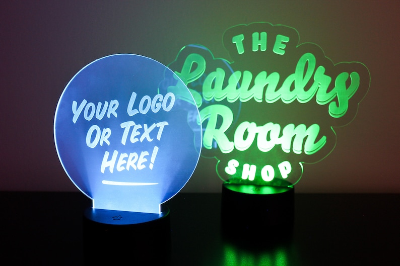 May include: Two illuminated LED signs. One is a blue circle with the text "YOUR LOGO OR TEXT HERE!". The other is a green sign that says "THE Laundry Room SHOP". Both signs are on black bases.