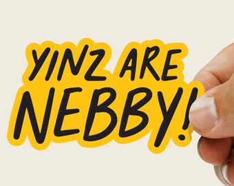 Op de afbeelding: Een gele sticker met de tekst "YINZ ARE NEBBY!" in het zwart, vastgehouden door een hand. De sticker heeft een afgeronde, onregelmatige vorm en een zwarte rand. De achtergrond is lichtbeige.