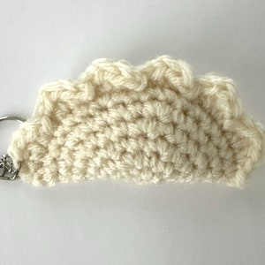 May include: Cream-colored, crocheted keychain in a half-circle shape. The keychain has a scalloped edge and a silver-colored metal ring with a small heart charm. The yarn appears to be a soft, textured material.