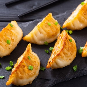 May include: Six golden brown dumplings with green onions on top, arranged on a black slate plate.