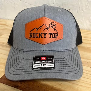 May include: Grey and black trucker cap with a leather patch displaying "Rocky Top" and a mountain design. The cap has a curved peak and an adjustable strap. The Richardson brand label is visible.
