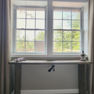May include: A wooden desk with a natural finish stands in front of a window. The desk has a rectangular top and two solid legs. A small birdhouse and a decorative item sit on the desk. A black power cord hangs from the desk.