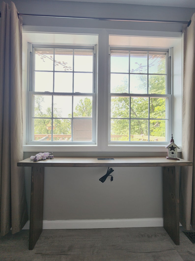May include: A wooden desk with a natural finish stands in front of a window. The desk has a rectangular top and two solid legs. A small birdhouse and a decorative item sit on the desk. A black power cord hangs from the desk.