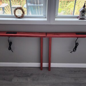 May include: Two red wooden console tables with angled legs. Each table has a rectangular top with a recessed area and a black power cord hanging down. The tables are positioned against a gray wall.