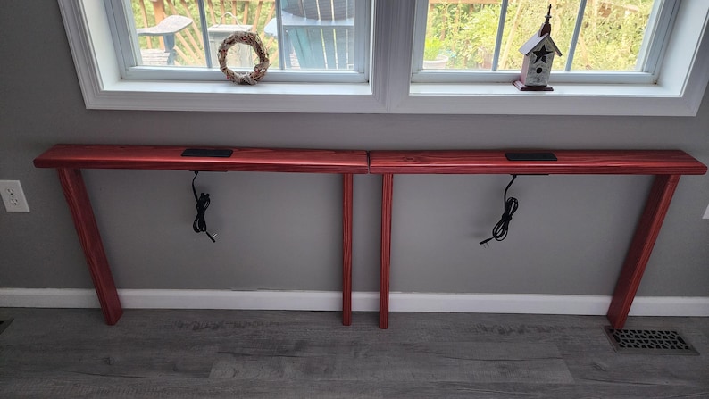 May include: Two red wooden console tables with angled legs. Each table has a rectangular top with a recessed area and a black power cord hanging down. The tables are positioned against a gray wall.