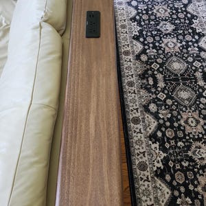 May include: A long, narrow, rectangular wooden console table with a smooth brown finish positioned behind a cream-colored sofa. The table features a black, rectangular electrical power outlet strip embedded flush into the center of the surface.