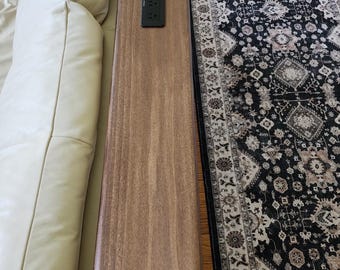 May include: A long, narrow, rectangular wooden console table with a smooth brown finish positioned behind a cream-colored sofa. The table features a black, rectangular electrical power outlet strip embedded flush into the center of the surface.