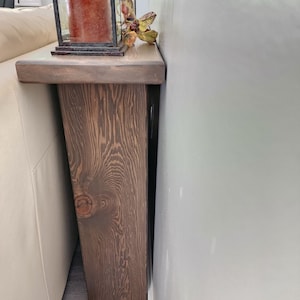 May include: A rustic, wooden sofa table with a dark brown finish. The table features a thick, rectangular top and a supporting leg with a knot detail. A decorative lantern with a candle and fall foliage sits on the table.