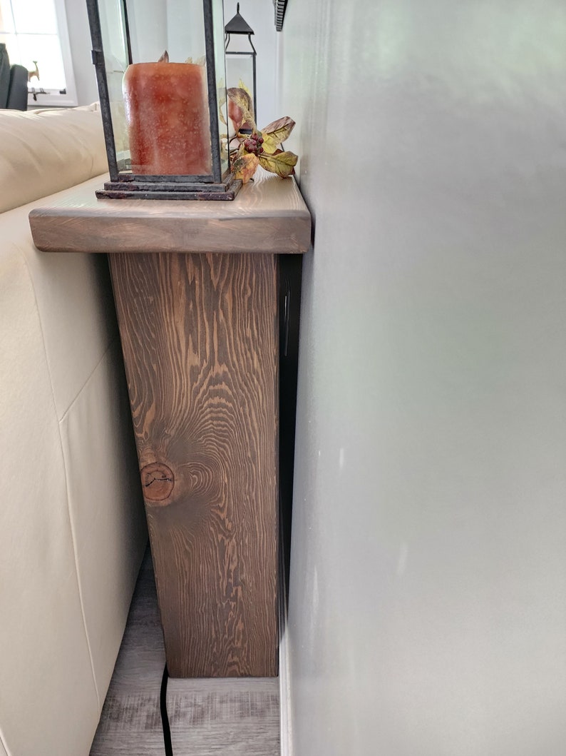 May include: A rustic, wooden sofa table with a dark brown finish. The table features a thick, rectangular top and a supporting leg with a knot detail. A decorative lantern with a candle and fall foliage sits on the table.