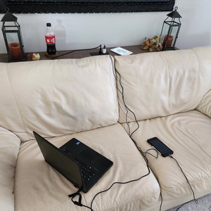 May include: A black laptop and a smartphone are connected to a power source via black charging cables, resting on a cream-colored leather sofa. A bottle of soda and decorative lanterns are visible in the background.