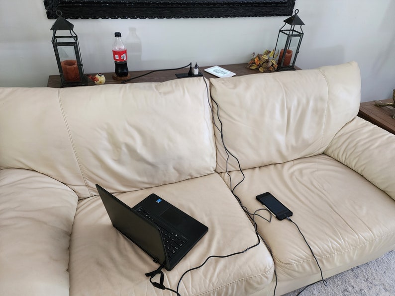 May include: A black laptop and a smartphone are connected to a power source via black charging cables, resting on a cream-colored leather sofa. A bottle of soda and decorative lanterns are visible in the background.