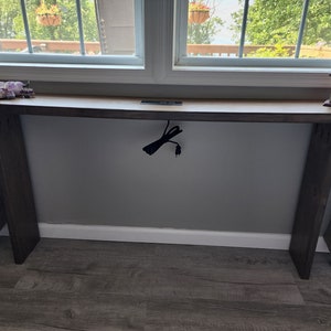 May include: A long, rectangular wooden console table with a natural wood top and dark brown legs. The table is positioned in front of a window. Decorative items, including crystals and a birdhouse, are placed on the table's surface.