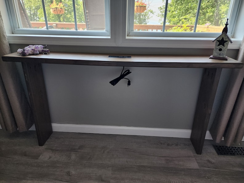 May include: A long, rectangular wooden console table with a natural wood top and dark brown legs. The table is positioned in front of a window. Decorative items, including crystals and a birdhouse, are placed on the table's surface.