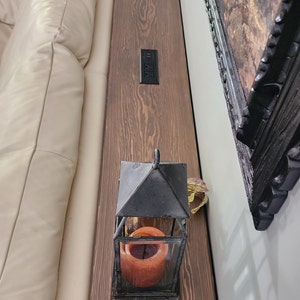 May include: A wooden sofa table with a dark stain, featuring a built-in power outlet. A black metal lantern with a lit orange candle sits on the table. A framed painting hangs on the wall.