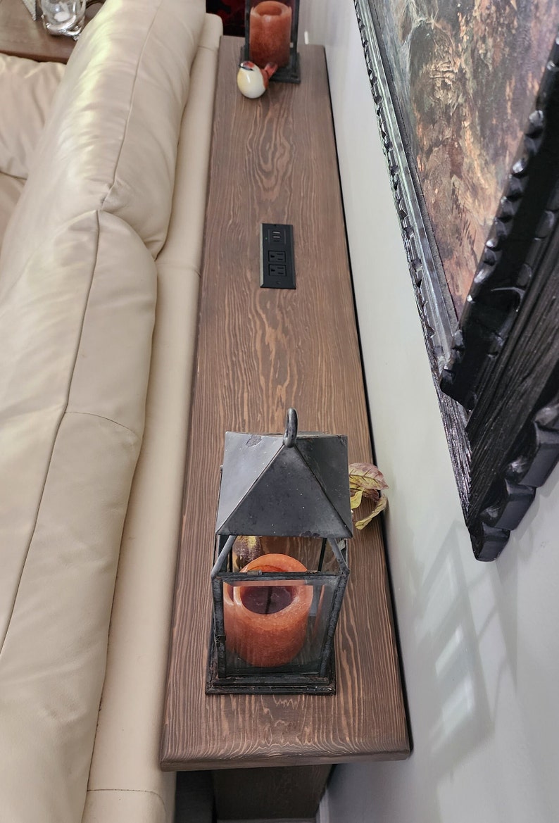 May include: A wooden sofa table with a dark stain, featuring a built-in power outlet. A black metal lantern with a lit orange candle sits on the table. A framed painting hangs on the wall.