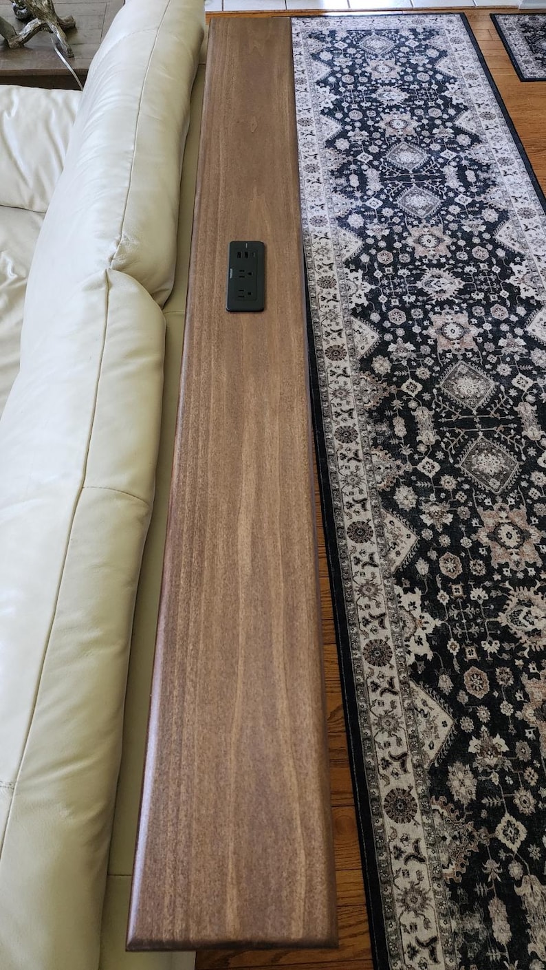 May include: A long, rectangular wooden table with a built-in electrical outlet. The table is a medium brown color and is positioned next to a cream-colored sofa. A patterned rug in black, white, and beige is visible on the floor.