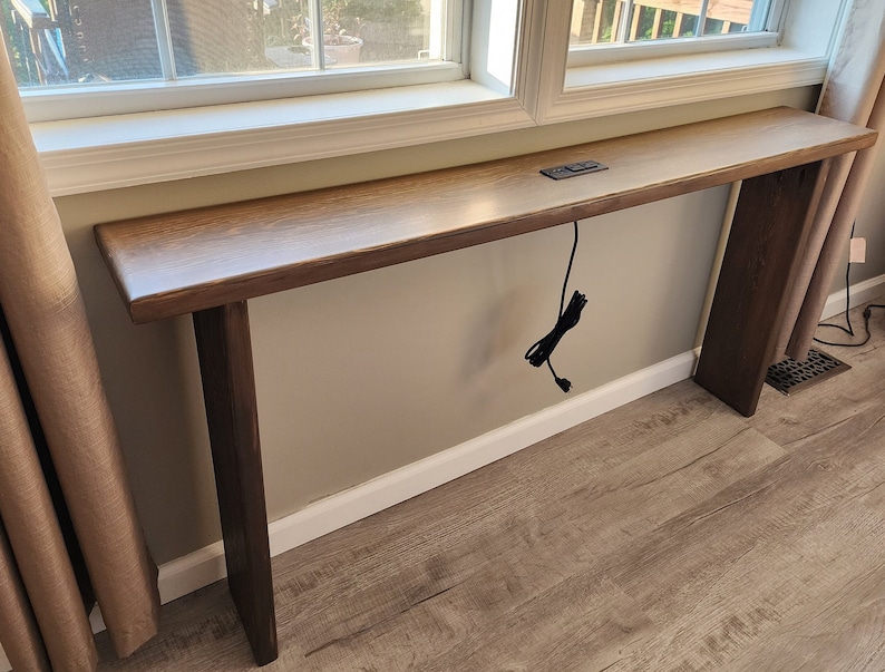 May include: A long, rectangular wooden console table with a light brown top and dark brown legs. The table is positioned in front of a window and has a power cord hanging down. A small remote is on the table.